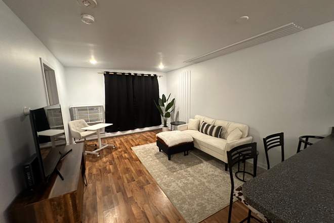 Open concept living room with TV, couch and portable/height adjustable desk. Sliding door with access to patio and natural light. - West Village 3