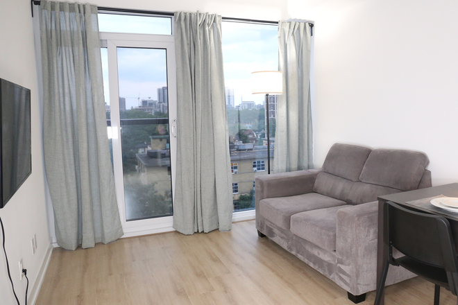 Large furnished living room with television, couch, curtains, floor to ceiling windows, and a sliding door with balcony railing. - 1 Bedroom Apartment Fully Furnished Turnkey Downtown