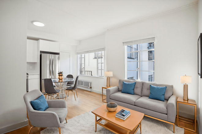 Living room with large window allowing natural light to flood the space, features a sofa. coffee table, dining table with chairs and a refrigerator - 2363 Le Conte Apartments