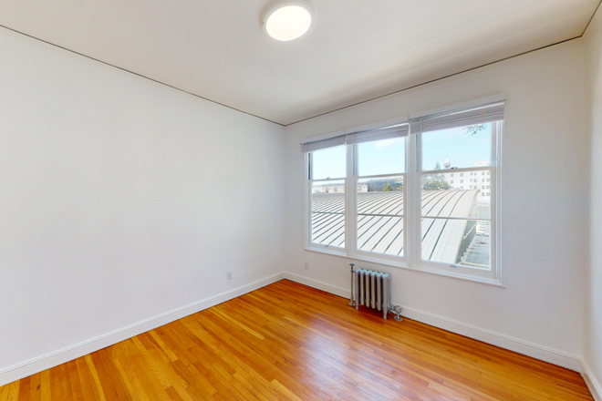 Small, empty room with wood flooring, large windows showcasing an outside view. A metal radiator sits beneath the window, creating a bright, open atmosphere. - 2605 Haste Street
